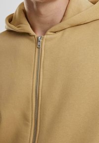 Beiger Kapuzenpullover mit weicher Textur, ausgestattet mit einer Kapuze und einem silbernen Metallreißverschluss, der die gesamte Vorderseite entlangläuft.