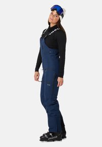 Navy blauwe skikleding met ritsen en zakken, gedragen over een zwart langmouwtop, gecombineerd met zwarte skischoenen en een skibril.