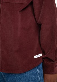 Camisa de manga larga de pana burdeos con un bolsillo en el pecho y una etiqueta cerca del dobladillo, combinada con jeans de denim azules. Tejido texturizado.