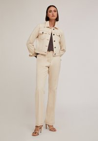 Veste en denim beige avec deux poches poitrine, associée à un pantalon taille haute assorti. Le mannequin porte des talons fins, mettant en valeur un haut texturé marron.