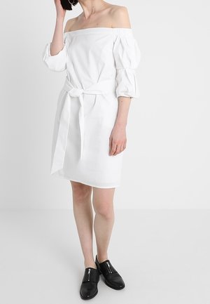 Femme portant une robe blanche épaules dénudées avec des manches bouffantes et une ceinture à la taille, associée à des chaussures noires sans lacets avec des fermetures éclair.