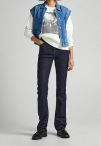 Gilet di jeans blu sopra un maglione bianco con design grafico nero, abbinato a jeans a campana blu scuro e scarpe nere con suole testurizzate.
