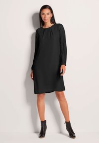 Schwarzes Langarmkleid mit Rundhalsausschnitt und plissierten Details an den Ärmeln; glatte Textur und knielanges Design. Getragen mit schwarzen Stiefeletten.