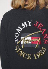 Schwarzes Langarmshirt mit einem kreisförmigen Logo auf der Rückseite in Weiß, Rot und Gold sowie Textakzenten "Tommy Jeans" und "Since 1985".