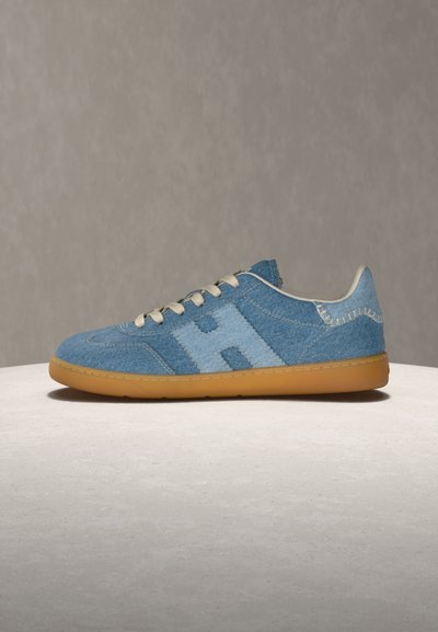Μπλε τζιν αθλητικό παπούτσι με ραφές "H" σε ανοιχτό μπλε, μπεζ κορδόνια και σόλα από καουτσούκ, τοποθετημένο σε ανοιχτόχρωμη επιφάνεια με απλό γκρι φόντο.