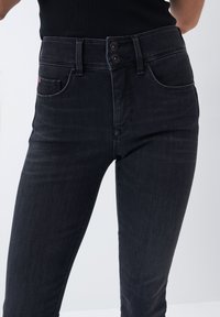 Jeans negros de cintura alta con ajuste slim, cierre de dos botones y bolsillos frontales. La textura desgastada en la tela añade un detalle sutil.