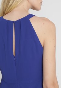 Femme portant une robe bleu roi sans manches avec un dos ajouré boutonné et une fermeture éclair, vue de dos et de côté.