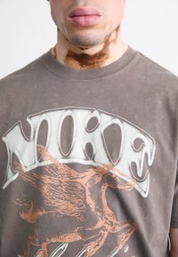 Brun bomulds t-shirt med hvid og kobber grafisk tryk af en vinget hest, korte ærmer, rund halsudskæring og en falmet tekstur.