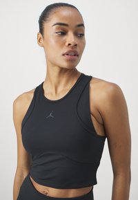 Top deportivo negro con un diseño ajustado y estilo de espalda nadadora. Presenta una cinturilla texturizada y un pequeño logo gris en la parte delantera.