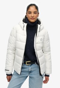 Hvid dynejakke med hætte, med et quiltet design, sort lynlås og logo på ærmet. Båret over en marineblå strikket sweater.