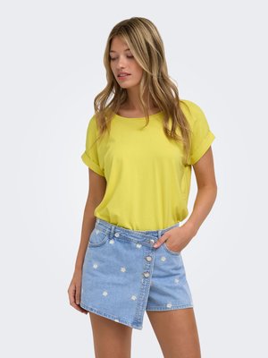 Femme portant un haut jaune à manches courtes et une jupe en jean asymétrique avec de petits imprimés floraux blancs, posant avec une main dans la poche.