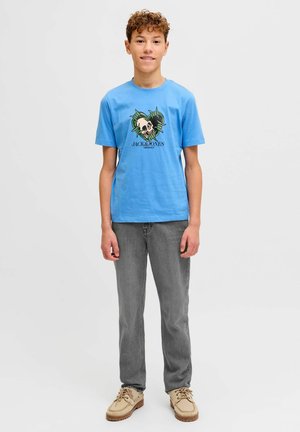 Light blue cotton t-shirt featuring a graphic skull design surrounded by green leaves. Worn with gray jeans and beige shoes.