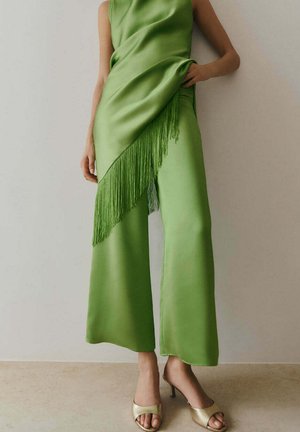 Vrouw draagt felgroene satijnen broek met bijpassende gesuikerde ceintuur, gecombineerd met gouden open-toe hakken, staand tegen een effen muur.