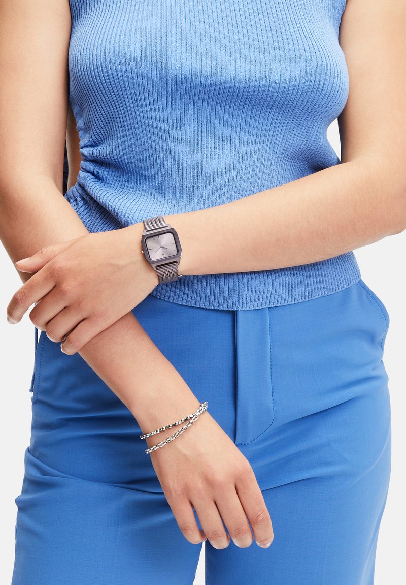 Vierkante horloge met een grijze metalen band, gecombineerd met een nauwsluitende blauwe ribgebreide top en blauwe broek. Armband met zilveren accenten om de pols.