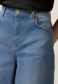 Jeans in denim blu chiaro con vita alta, chiusura con bottone argento e dettagli di tasche laterali. La cucitura è visibile lungo le cuciture.