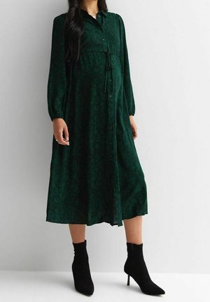 Frau trägt ein dunkelgrünes Midi-Kleid mit langen Ärmeln und Knopfleiste vorne, mit dezentem Blumenmuster sowie schwarze Stiefeletten mit Absatz.