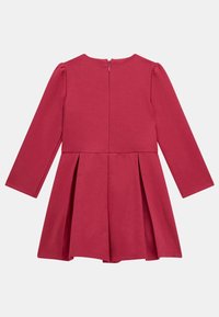 Robe à manches longues en rose uni. Présente un col rond, une fermeture éclair au dos et une jupe plissée. Confectionnée en tissu doux et extensible.