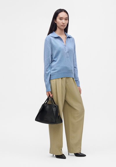 Femme portant un pull bleu clair et un pantalon large beige, tenant un sac à main noir et portant des talons pointus noirs.