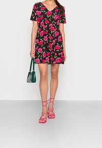 Robe courte à fleurs avec un fond noir, présentant des fleurs vives roses et vertes, associée à des sandales à brides roses et un sac à main vert.