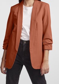 Rostfärgad kavaj i ett mjukt tyg med avslappnad passform, sjalkrage och upprullade ärmar, kombinerad med en vit t-shirt och mörk jeans.