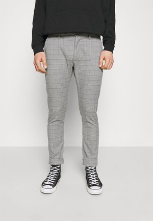 Mężczyzna ubrany w szare, wąskie spodnie w kratę ze zrolowanymi mankietami oraz czarne sneakersy za kostkę z białymi sznurówkami, widoczny czarny sweter.
