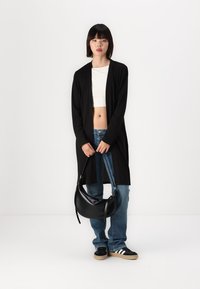Black long cardigan made of lightweight fabric, worn over a white crop top. Paired with blue jeans and a black curved handbag.
