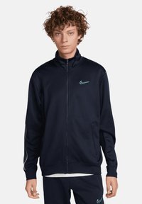 Chaqueta deportiva de navy con cierre y cuello alto, logo de Nike en azul claro, bolsillos laterales y detalles blancos en las mangas. Tejido suave y texturizado.