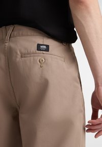 Pantaloni beige in tessuto liscio, dotati di una tasca posteriore con chiusura a bottone e un'etichetta nera del logo Vans sopra la tasca.