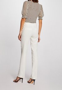 Pantalones blancos ajustados de cintura alta con una textura suave. Combinados con una blusa transparente y estampada que presenta mangas abullonadas y un diseño geométrico.