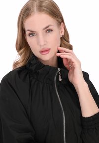 Fekete bélelt dzseki magas fodros gallérral, ezüst cipzárral és rugalmas mandzsettákkal; sima tapintású, minimalista dizájn.