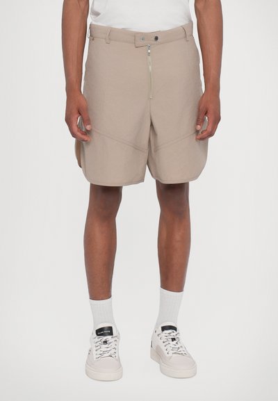 Beige Shorts mit einem Reißverschluss und einem Knopfverschluss, zwei Seitentaschen, diagonalen Nähten und einer entspannten Passform. Getragen mit weißen Socken und Turnschuhen.