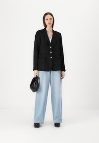 Blazer noir texturé avec deux poches avant, trois boutons brillants, associé à un jean large bleu clair et un sac à main noir.