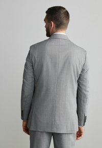 Chaqueta de traje gris con un acabado texturizado. Cuenta con una abertura trasera única y detalle de puño con tres botones. Camisa blanca visible en el cuello.