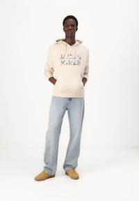 Haut à capuche beige avec un logo graphique coloré, associé à un jean bleu clair. La texture est douce et le jean a une coupe décontractée. Baskets marron.