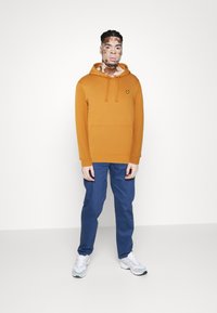 Sweatshirt à capuche orange avec une poche avant et un petit logo, assorti à un pantalon bleu et des baskets blanches, se tenant sur un fond blanc.