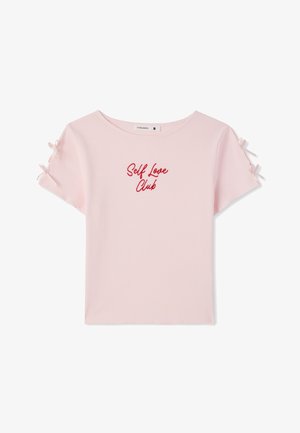 Maglietta rosa chiaro a maniche corte con motivo a costine, testo rosso "Self Love Club" centrato e piccoli fiocchi legati su ogni manica.