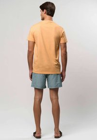 Férfi háttal áll, világos háttér előtt, barack színű, rövid ujjú pólót, kék csíkos rövidnadrágot és sötét szandált visel.