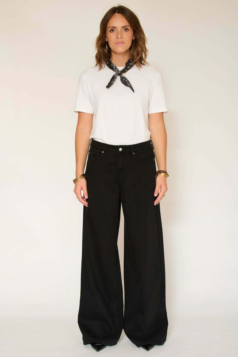 Jean noir à jambes larges et taille haute, associé à un t-shirt blanc uni. Un foulard noir à motifs est noué autour du cou pour accentuer la tenue.