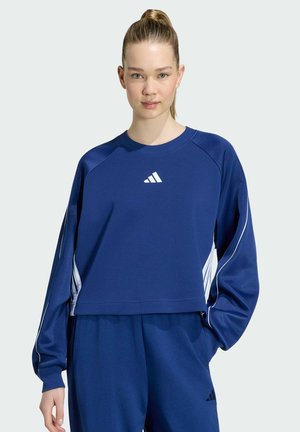 Junge Frau mit blondem Pferdeschwanz, die einen blauen Adidas-Trainingsanzug mit weißen Streifen trägt und mit einer Hand in der Tasche steht.