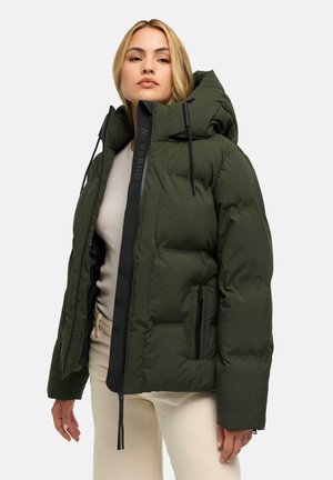 Frau trägt eine dunkelgrüne Steppjacke mit großer Kapuze, grauen Pullover und hellbeiger Hose, steht vor neutralem Hintergrund.