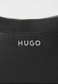 Schwarze, strukturiertes Lederhandtasche mit einem silbernen Reißverschluss. Besitzt ein auffälliges, metallisches "HUGO"-Logo auf der Vorderseite.