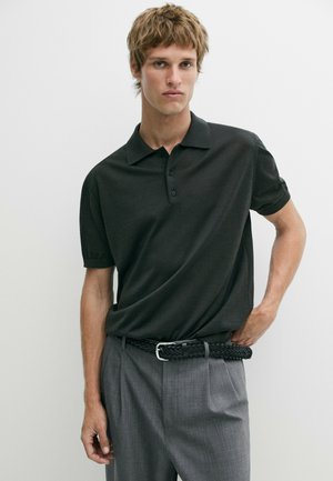 Junger Mann steht vor einem schlichten Hintergrund, trägt ein schwarzes Poloshirt, graue Hosen und einen schwarzen geflochtenen Gürtel und schaut in die Kamera.