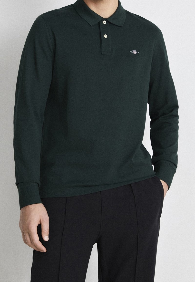 Long-sleeve dark green polo shirt with a soft texture, two-button placket, and a small logo on the chest. Paired with dark trousers.