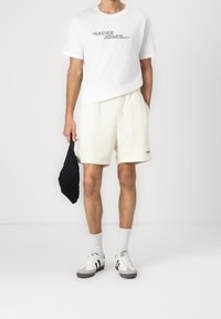 Weißes T-Shirt mit grauem Logo, cremefarbene Shorts, schwarze Tasche, weiße Socken und hellgraue Sneakers mit schwarzen Streifen. Einfaches, lässiges Outfit.