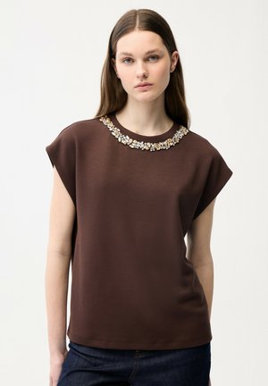 Jonge vrouw met lang bruin haar draagt een mouwloos bruin topje versierd met een kralenbloemmotief bij de hals, staand tegen een effen witte achtergrond.