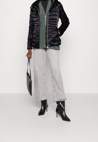 Veste matelassée noire avec épaules texturées, associée à un haut en maille vert, un jean gris clair à jambes larges, et des bottines à talons pointus noires.