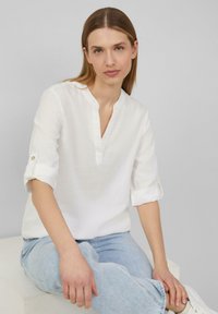 Blouse en lin blanc avec un col en V et des manches trois quarts, avec des poignets retroussés à boutons. Associée à un jean bleu clair, offrant une coupe décontractée.
