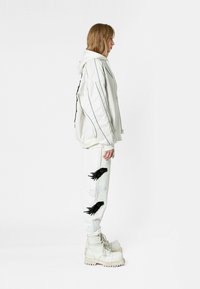 Chaqueta blanca con capucha y franjas negras, combinada con pantalones de chándal blancos con bordados de plumas en blanco y negro, y botas gruesas beige.