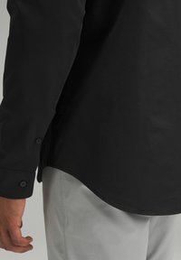 Maglietta nera a maniche lunghe con orlo curvo, polsini abbottonati e texture liscia. Indossata con pantaloni grigi chiari, visibile da una vista laterale parziale.