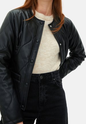 Kobieta w czarnej pikowanej skórzanej kurtce na beżowym swetrze oraz czarnych dżinsach z wysokim stanem, stoi z jedną ręką na biodrze.